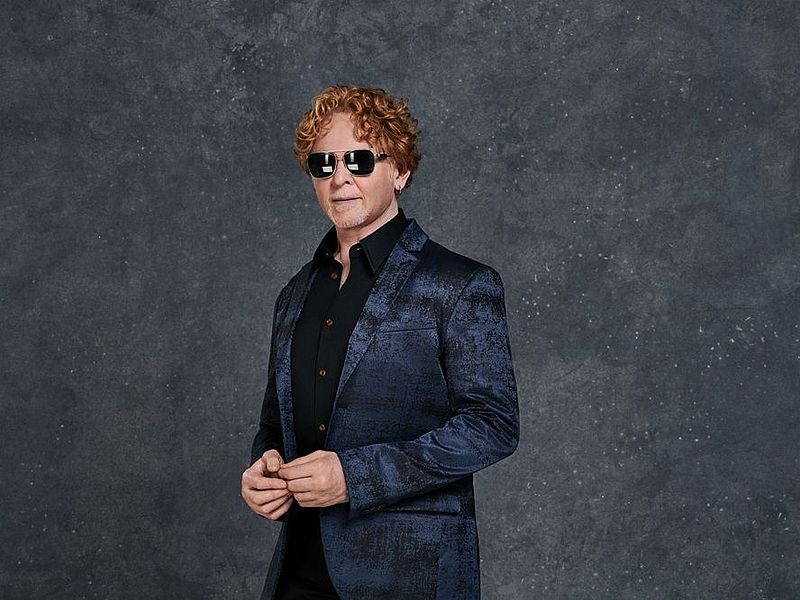Mick Hucknall steht vor einem grauen Hintergrund. Er trägt ein dunkelblaues Jacket und ein schwarzes Hemd. Er hat eine dunkle, reflektierende Sonnenbrille auf. 