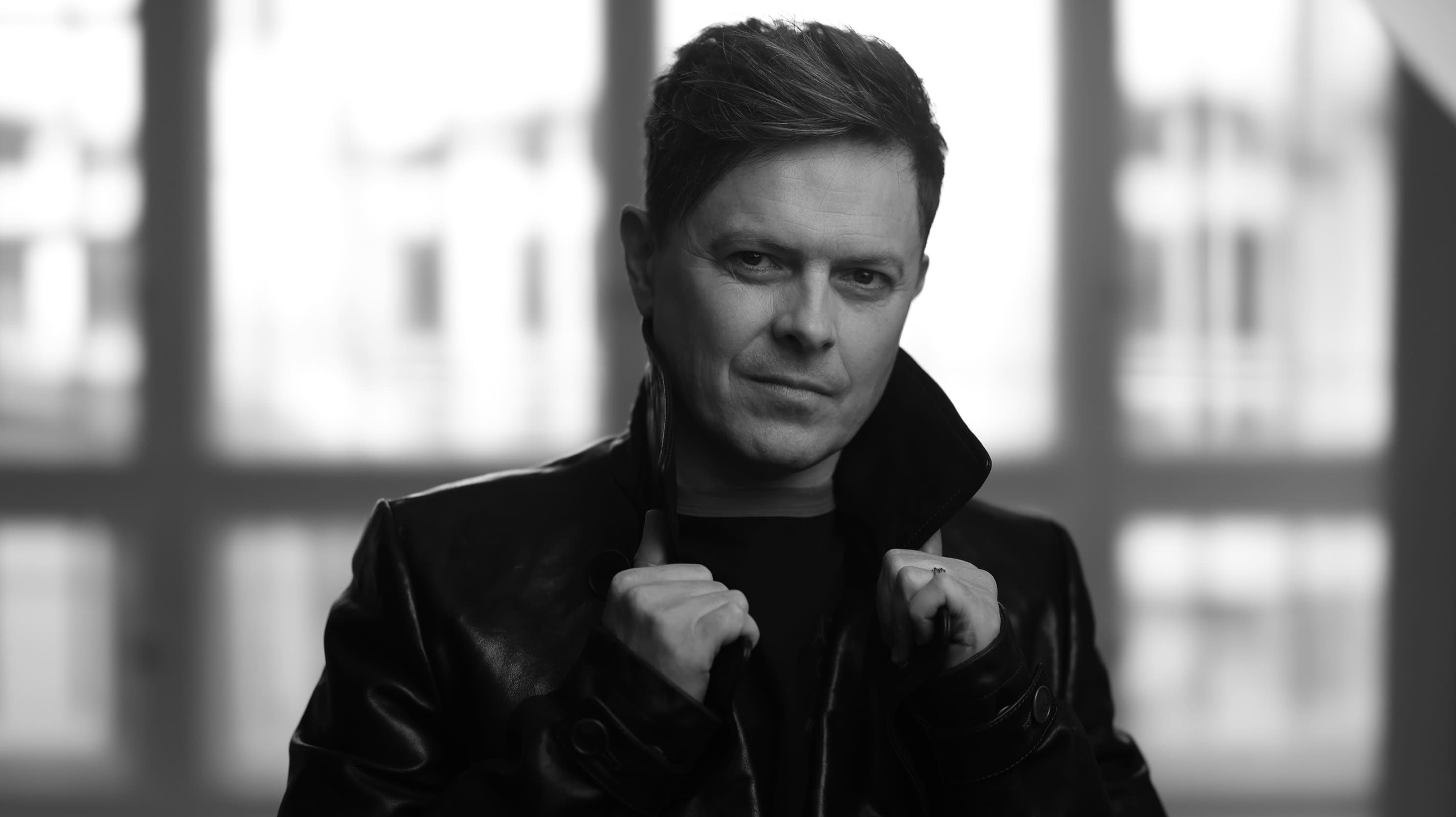 Michael P. Kelly trägt eine Lederjacke, hat die Hände am Kragen. Er steht vor einem großen Fenster. 