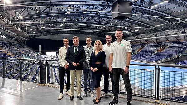 Sechs Personen stehen in der leeren, hell beleuchteten Multifunktionshalle. Im Hintergrund ist das Handball-Parkett. 