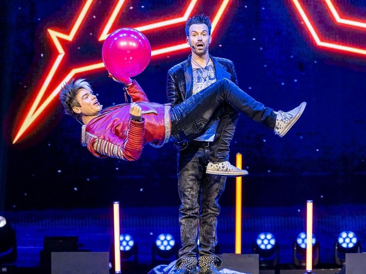 Die Ehrlich Brothers tragen beide Lederjacke und Jeans. Chris Ehrlich schwebt in der Luft und hält einen pinken Luftballon in der Hand. 