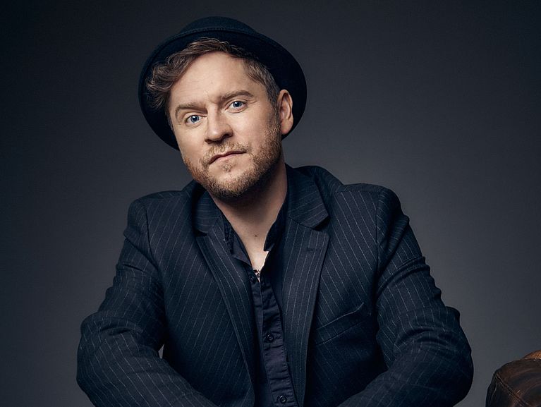 Johannes Oerding sitzt auf einem braunen Ledersessel. Er trägt einen schwarzen, gestreiften Anzug und einen Hut. 