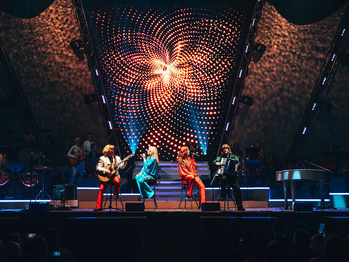 Zwei Männer und zwei Frauen sitzen auf Barhockern. Die Frauen singen, ein Mann hat eine Gitarre, der andere ein Akkordeon in der Hand. 