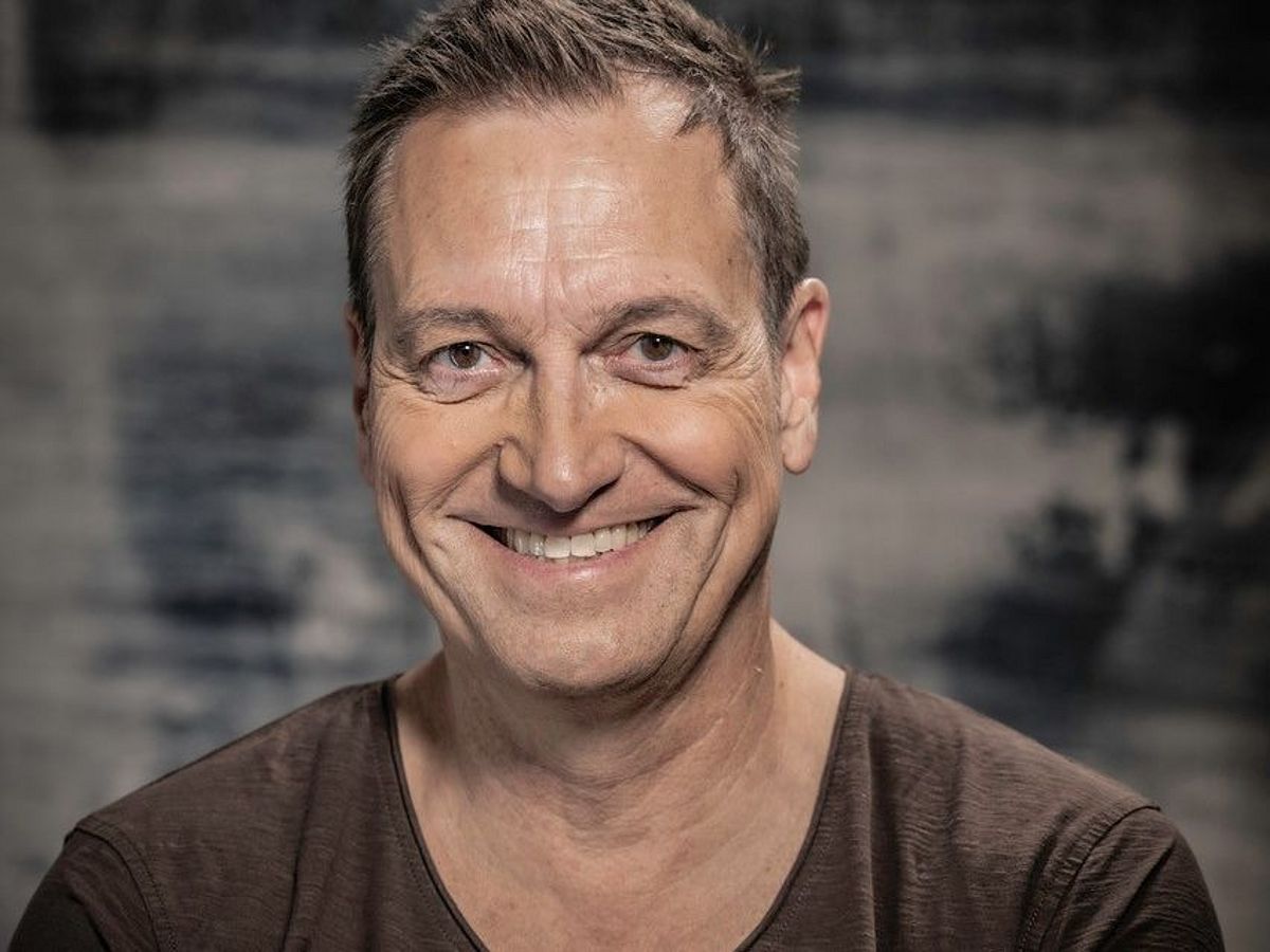 Pressefoto von Dieter Nuhr. Er trägt ein dunkles T-Shirt, verschränkt die Arme und lächelt. 