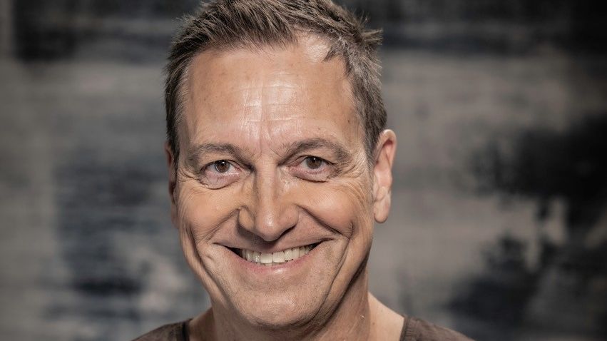 Pressefoto von Dieter Nuhr. Er trägt ein dunkles T-Shirt, verschränkt die Arme und lächelt. 