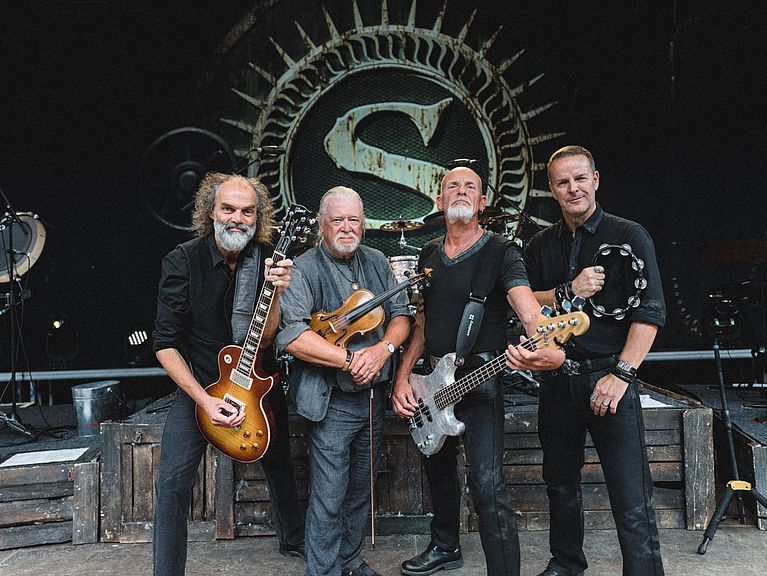 Die Band steht mit ihren Instrumenten auf der Bühne. Im Hintergrund ist ein großes S zu sehen. 