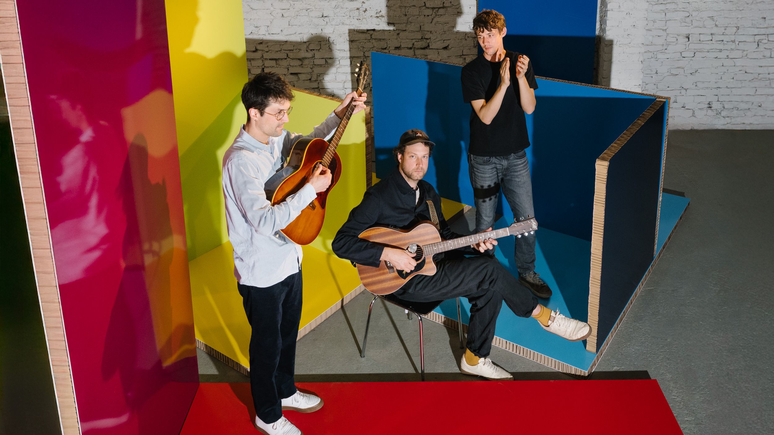 Pressefoto der Band AnnenMayKantereit. Zwei der Mitglieder stehen, einer sitzt auf einem Stuhl. Zwei haben eine Gitarre in der Hand. Sie stehen in einer Halle, um sie herum sind Raumteiler in den Farben gelb, blau und rot aufgestellt. 