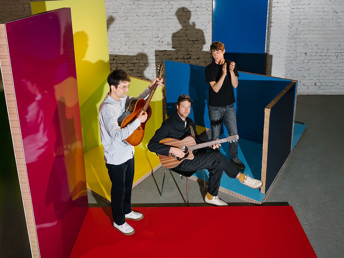 Pressefoto der Band AnnenMayKantereit. Zwei der Mitglieder stehen, einer sitzt auf einem Stuhl. Zwei haben eine Gitarre in der Hand. Sie stehen in einer Halle, um sie herum sind Raumteiler in den Farben gelb, blau und rot aufgestellt. 