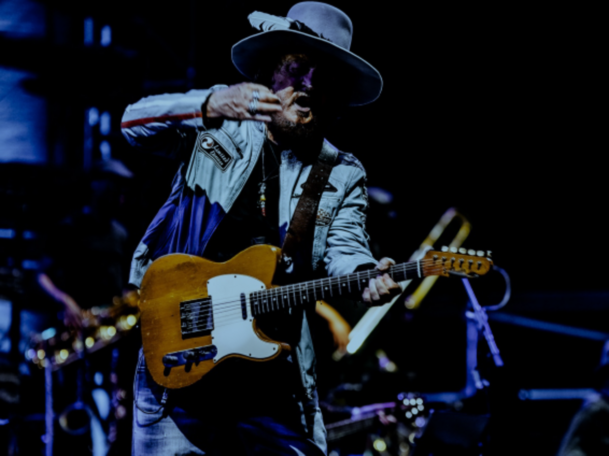 Zucchero steht mit E-Gitarre auf der Bühne. Er trägt einen Hut. Das Bild ist dunkel gehalten. 