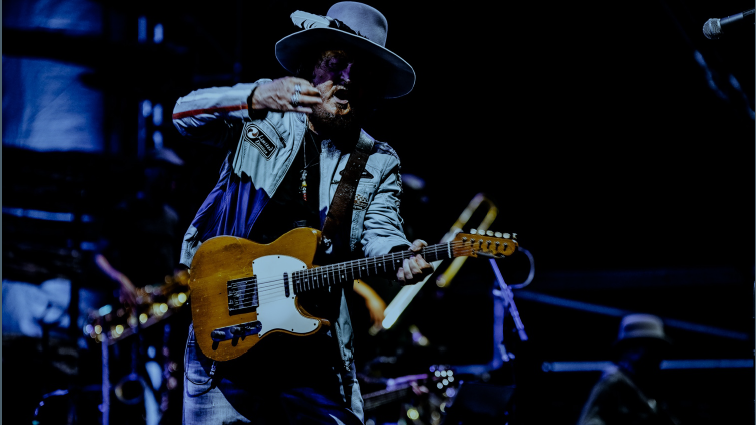 Zucchero steht mit E-Gitarre auf der Bühne. Er trägt einen Hut. Das Bild ist dunkel gehalten. 