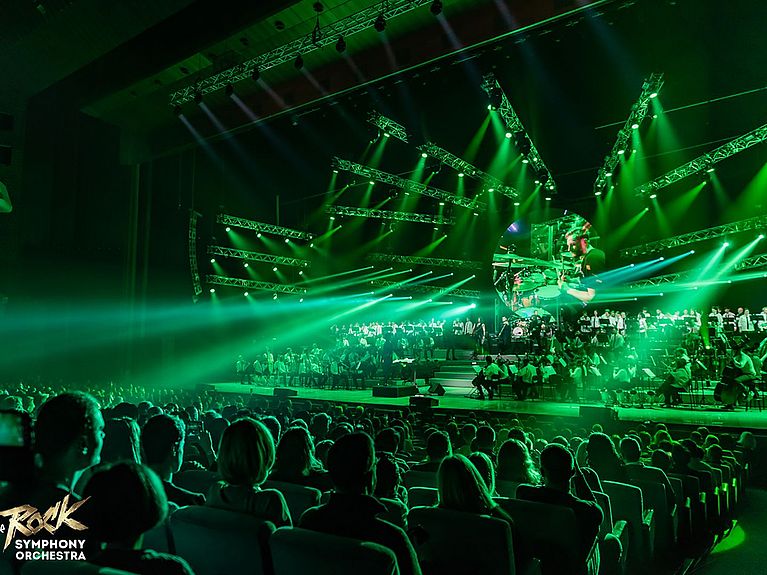 Ein Bild von einer Show. Blick vom Publikum auf die Bühne. Auf der Bühne ist ein großes Orchester. Die Szene erscheint im grünen Licht. 