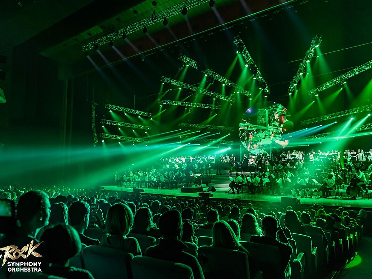 Ein Bild von einer Show. Blick vom Publikum auf die Bühne. Auf der Bühne ist ein großes Orchester. Die Szene erscheint im grünen Licht. 