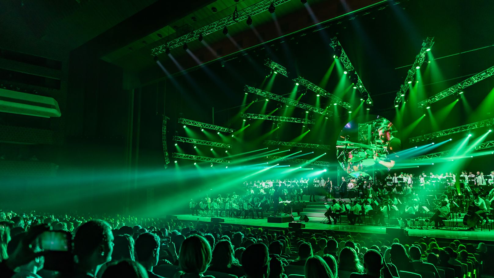 Ein Bild von einer Show. Blick vom Publikum auf die Bühne. Auf der Bühne ist ein großes Orchester. Die Szene erscheint im grünen Licht. 