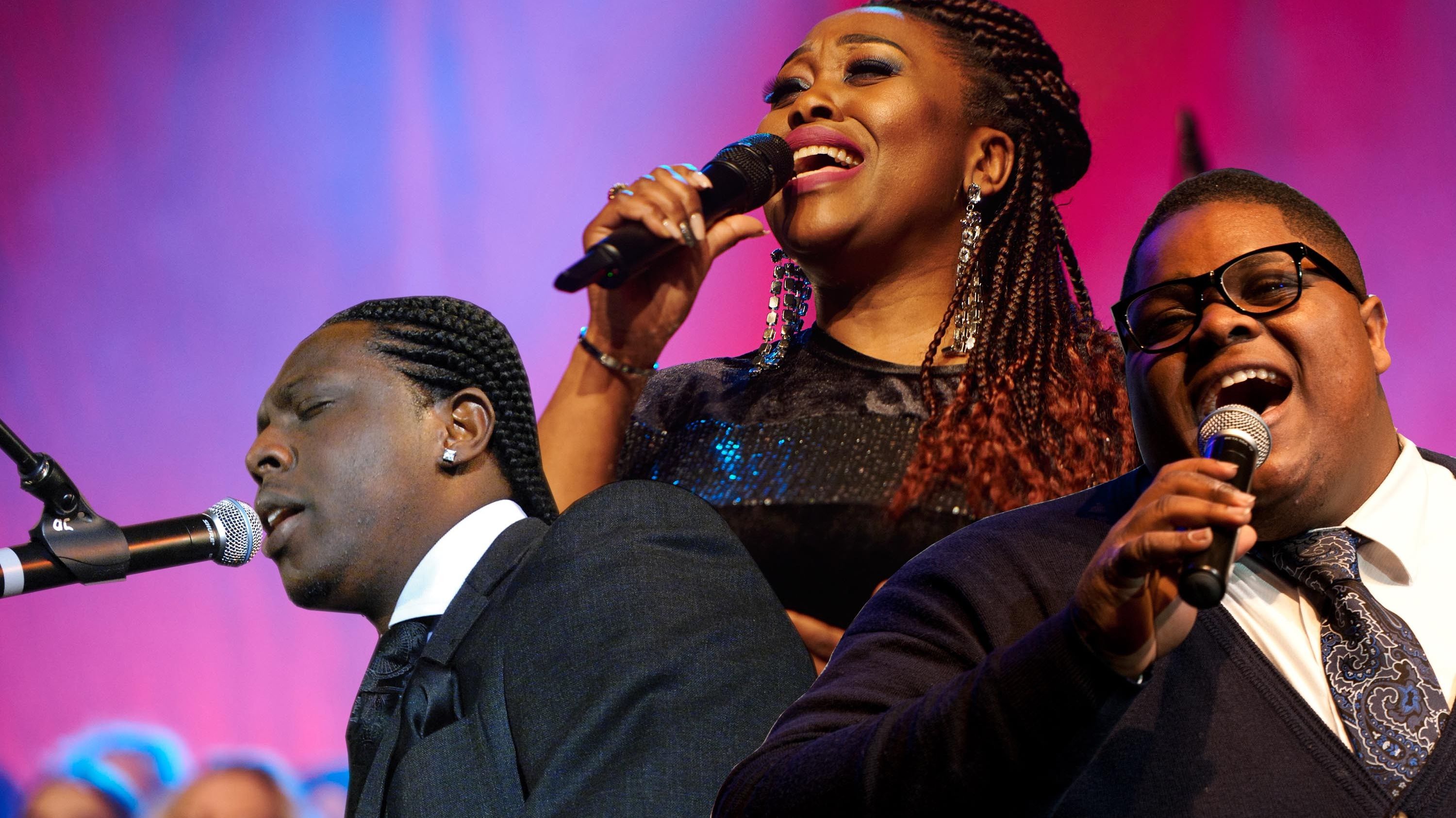 zwei Sänger und eine Sängerin jeweils mit Mikrofon sind zu sehen. 