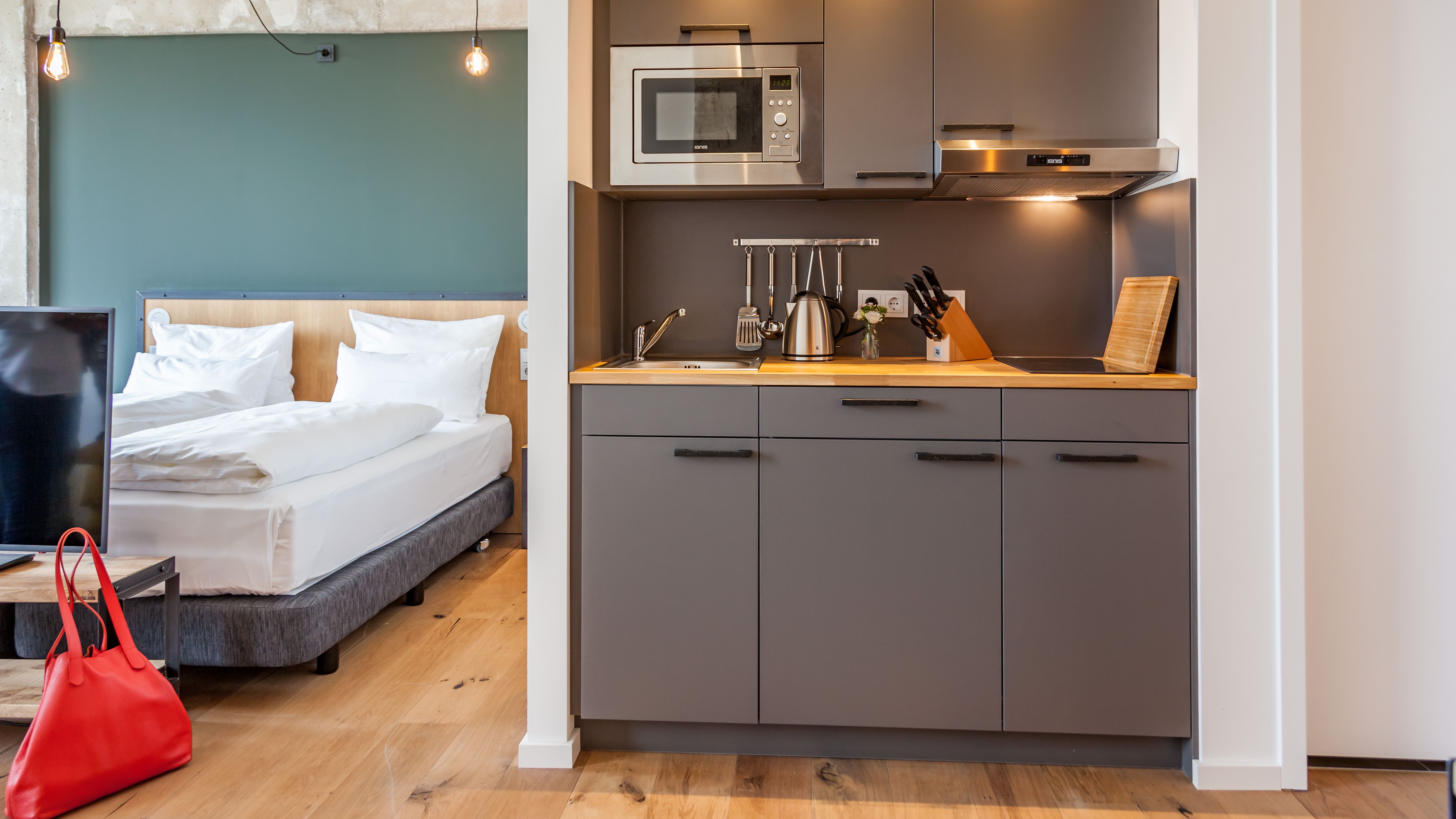 Blick in ein Hotelzimmer mit einer Küchenzeile. Vor dem Doppelbett steht ein Fernseher. Neben der Fernsehkommode steht eine rote Tasche. 