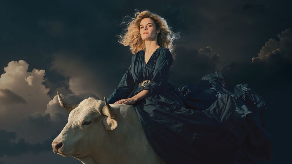 Hazel Brugger sitzt in einem blauen Kleid auf einem weißen Bullen, im Hintergrund sind Wolken zu sehen. Ihre langen blonden, lockigen Haare und ihr Kleid wehen im Wind. 