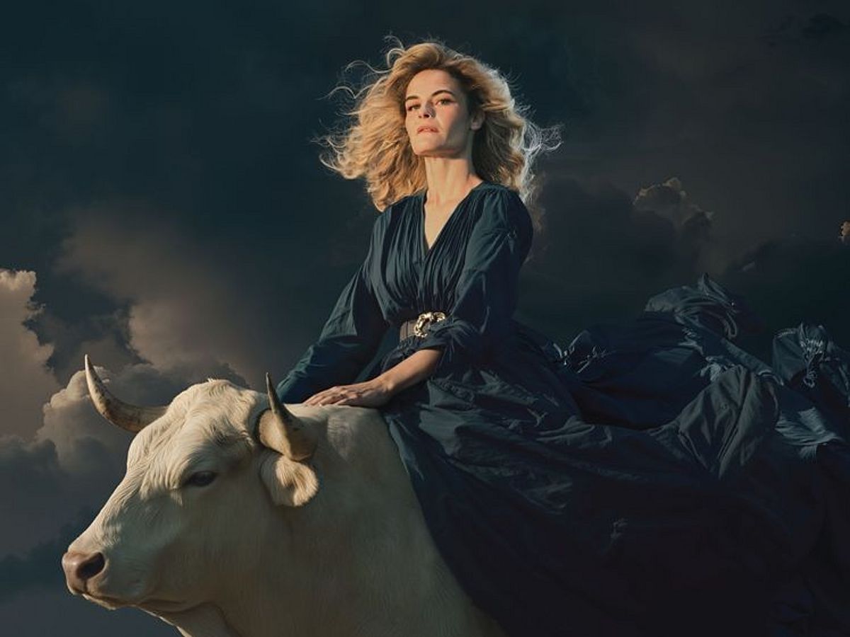 Hazel Brugger sitzt in einem blauen Kleid auf einem weißen Bullen, im Hintergrund sind Wolken zu sehen. Ihre langen blonden, lockigen Haare und ihr Kleid wehen im Wind. 