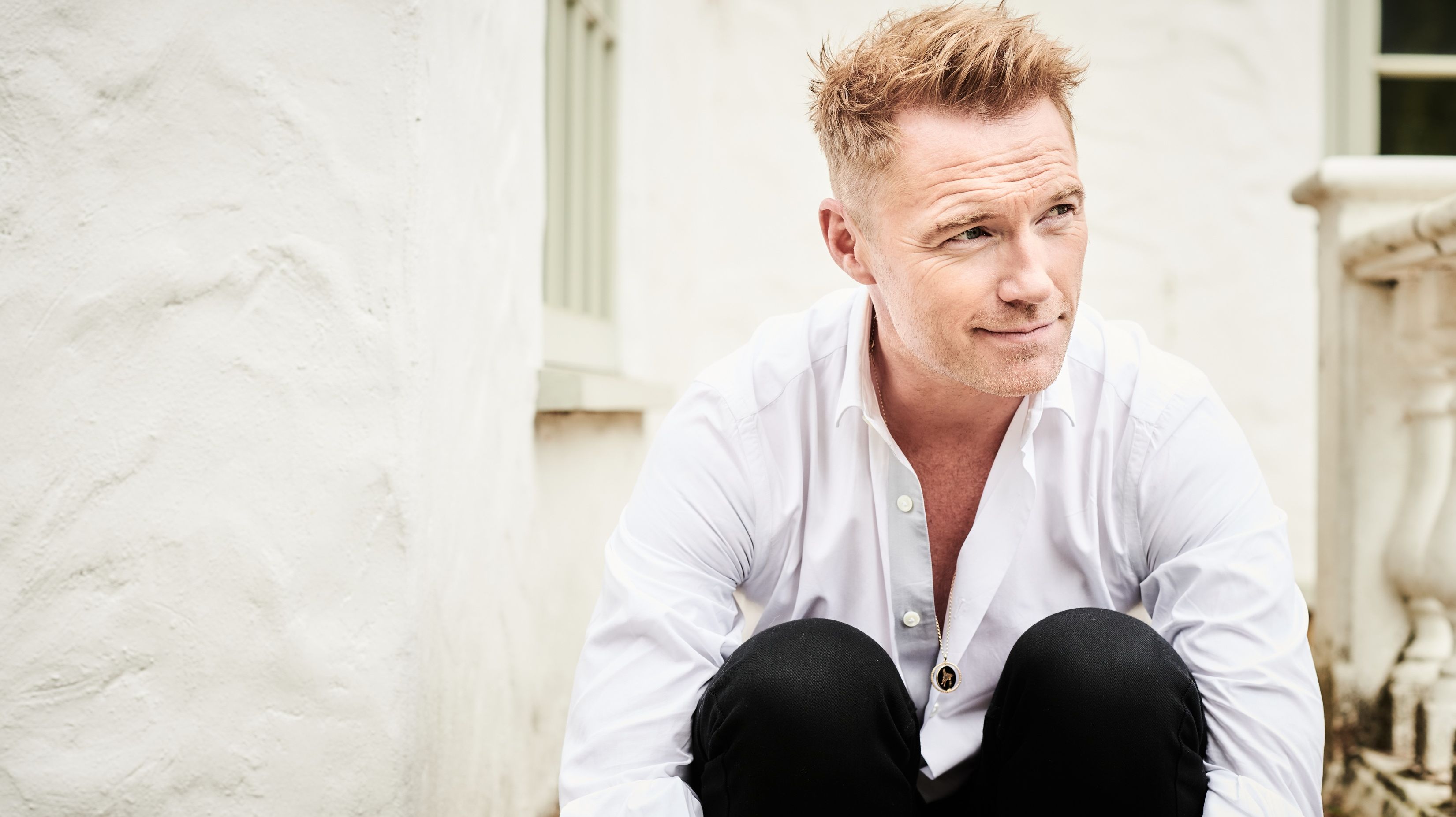 Ronan Keating sitzt vor einem weißen Gebäude auf einer Treppe. Er schaut nach rechts, runzelt die Stirn und umarmt seine Knie. Er trägt ein helles, aufgeknöpftes Hemd und eine schwarze Hose. 
