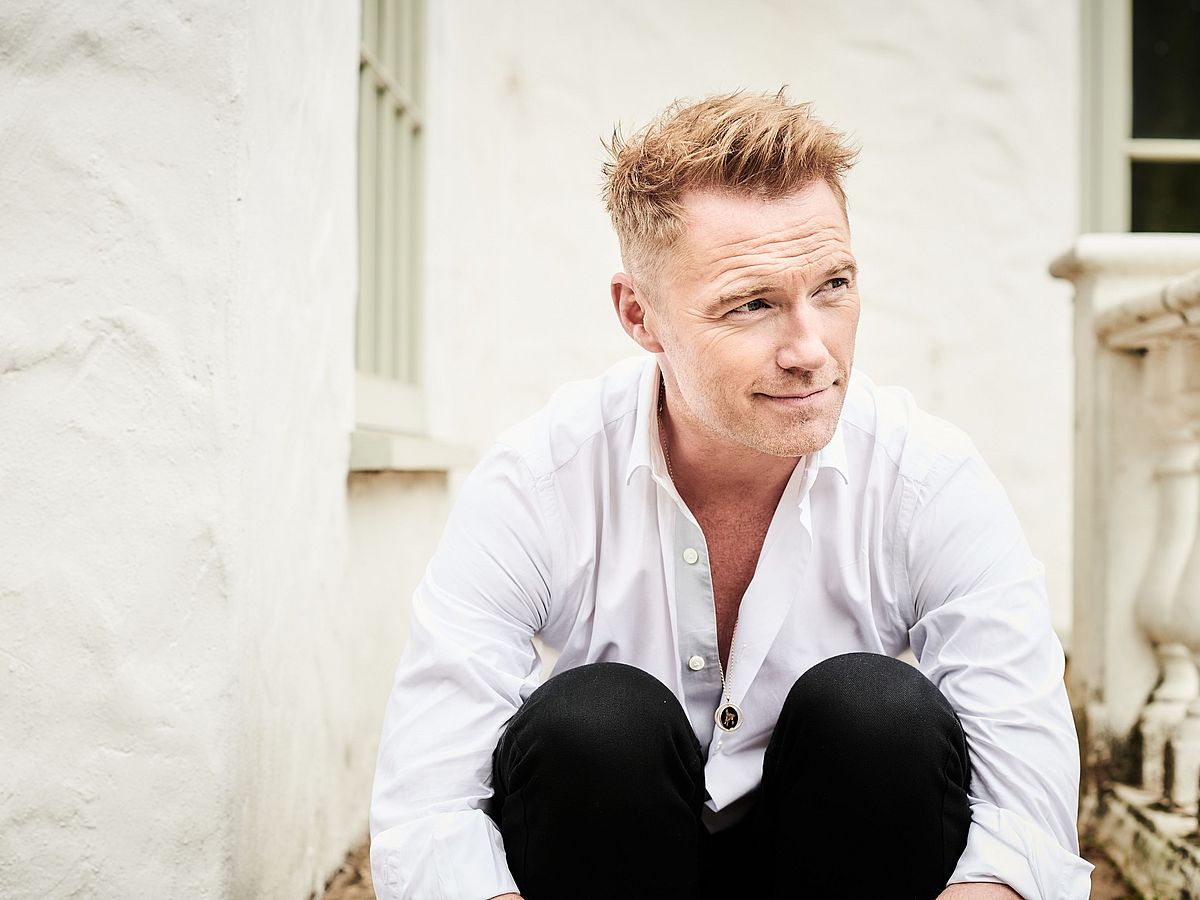 Ronan Keating sitzt vor einem weißen Gebäude auf einer Treppe. Er schaut nach rechts, runzelt die Stirn und umarmt seine Knie. Er trägt ein helles, aufgeknöpftes Hemd und eine schwarze Hose. 