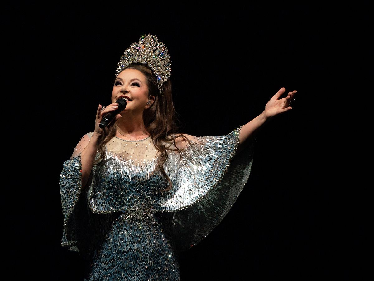 Vor einem schwarzen Hintergrund steht Sarah Brightman mit einem Mikrofon in der Hand. Sie trägt ein Abendkleid, das glitzert. In ihren Haaren hat sie große Tiara. 