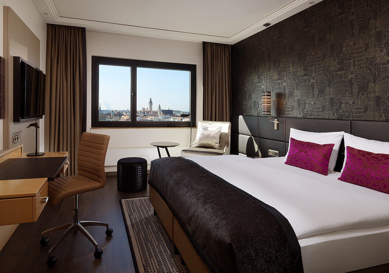 Ein modernes Hotelzimmer mit TV und Schreibtisch samt Stuhl auf der linken Seite und großem Doppelbett hinter dem eine dunkle Tapete ist auf der rechten Seite.