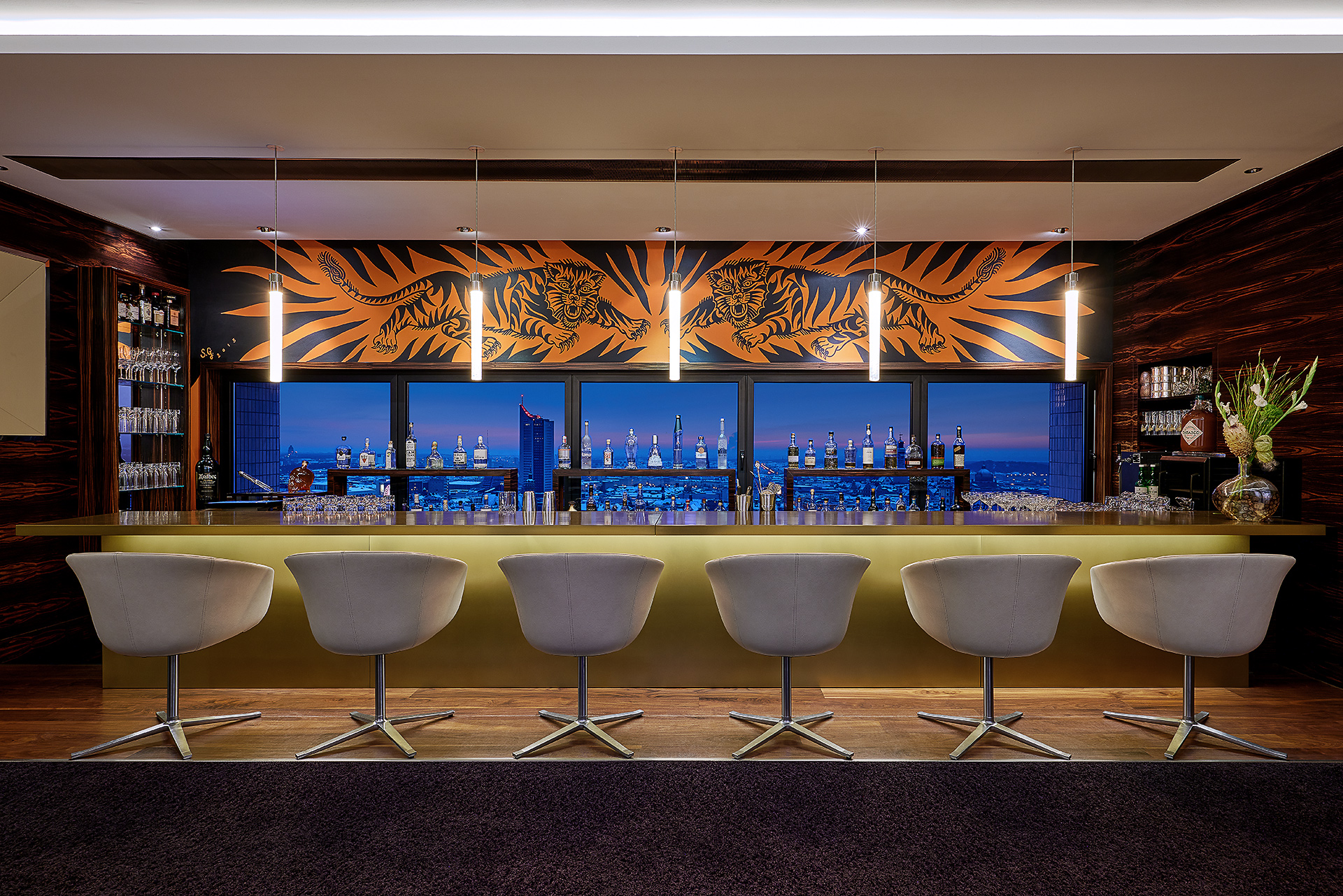 Blick auf sechs halbhohe Stühle die an einer eleganten Bar stehen mit Blick auf die Leipziger Skyline