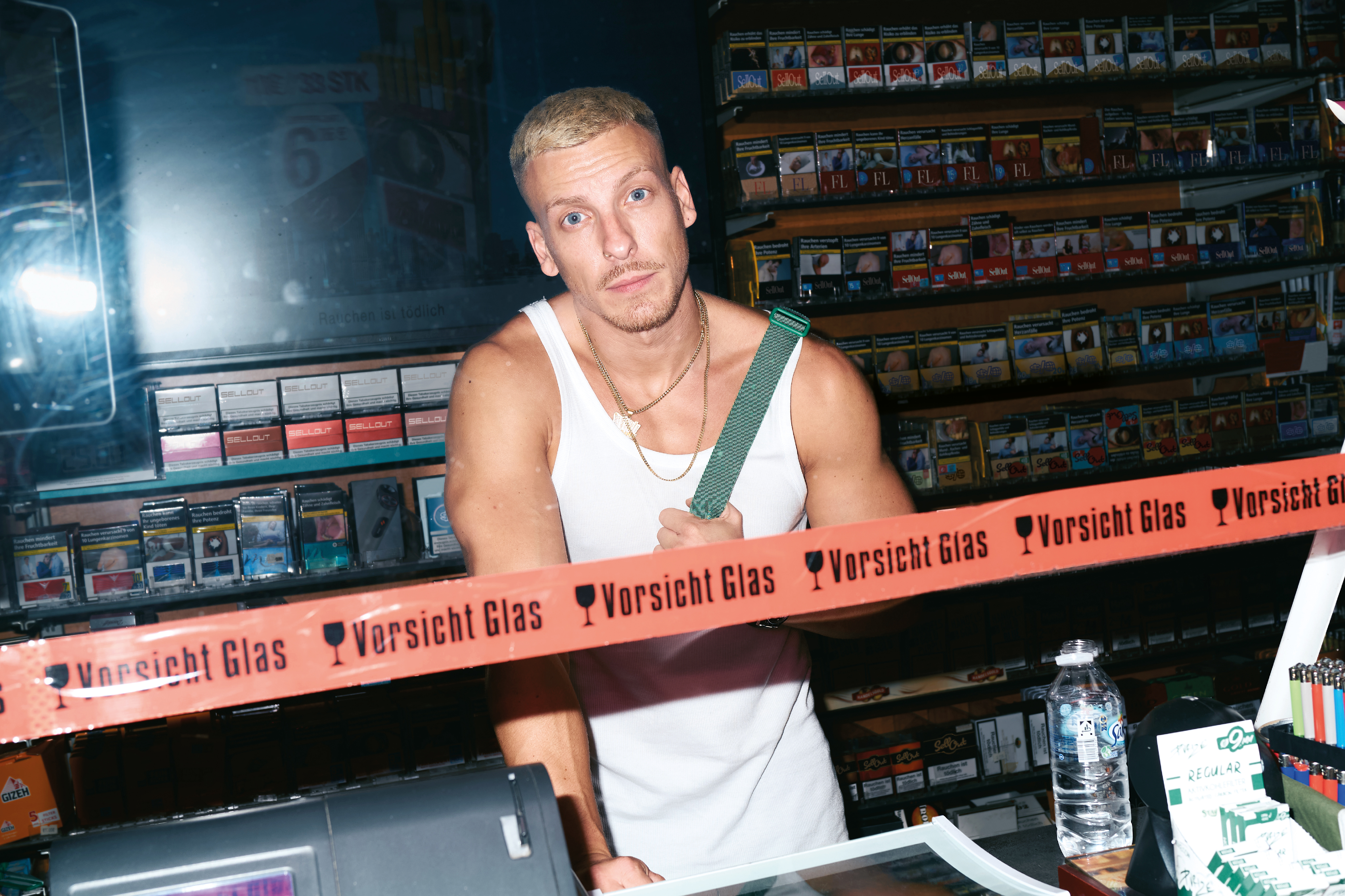 Felix Lobrecht hat ein Unterhemd mit Kette an und steht hinter einem Kiosk-Tresen. 