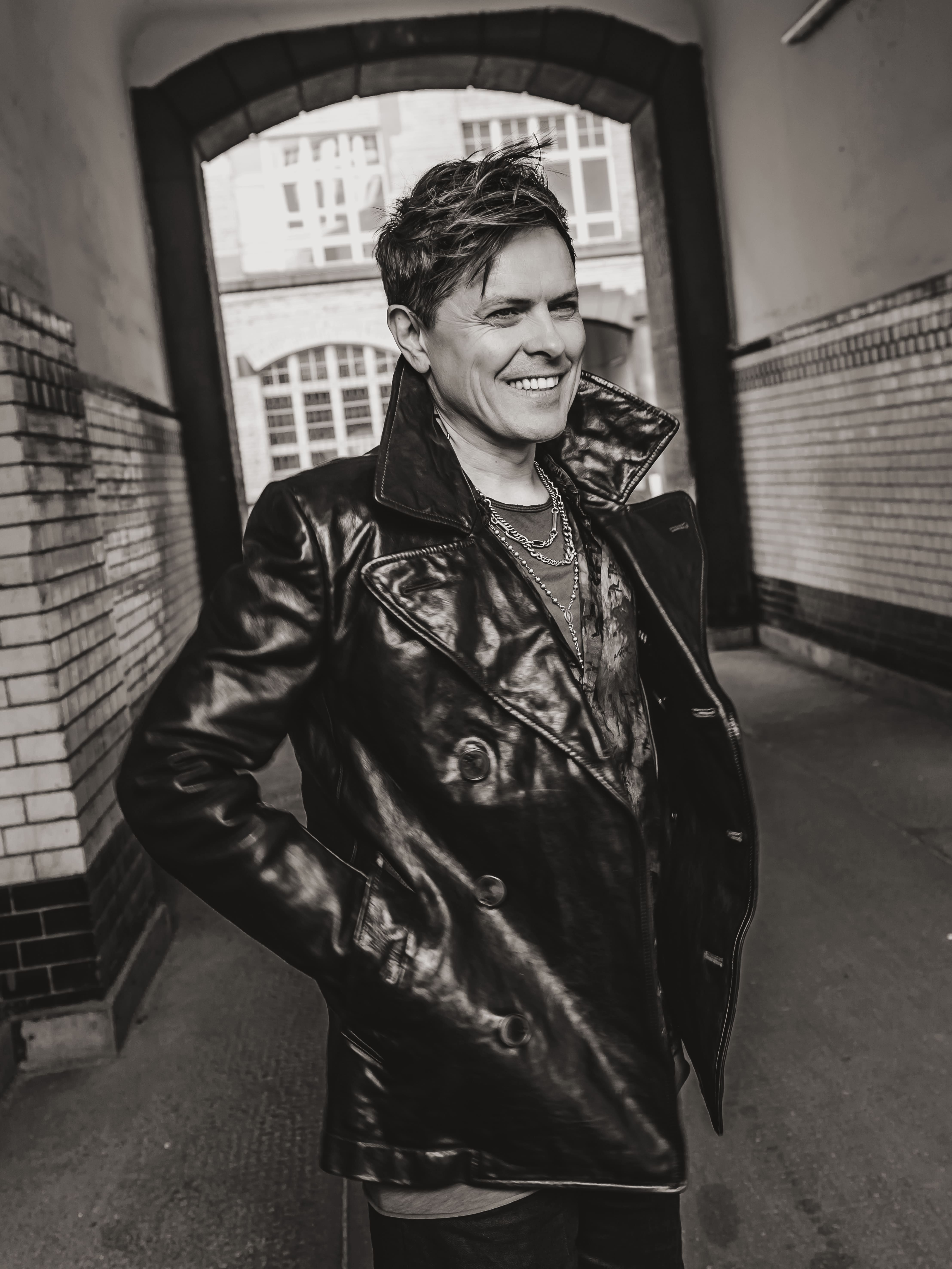 Michael P. Kelly hat eine Lederjacke an. Er steht in einer Hofeinfahrt. 