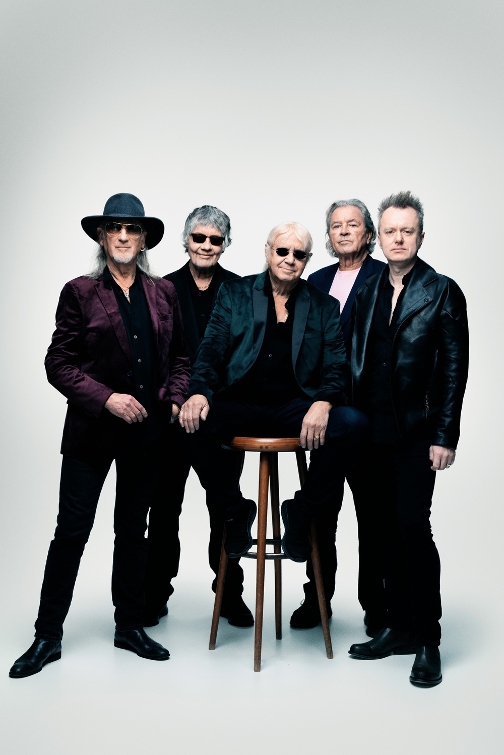 Bandfoto von Deep Purple vor einem weißen Hintergrund. Alle tragen dunkle Kleidung, drei von ihnen haben eine Sonnenbrille an. Einer von ihnen sitzt auf einem Barhocker. 