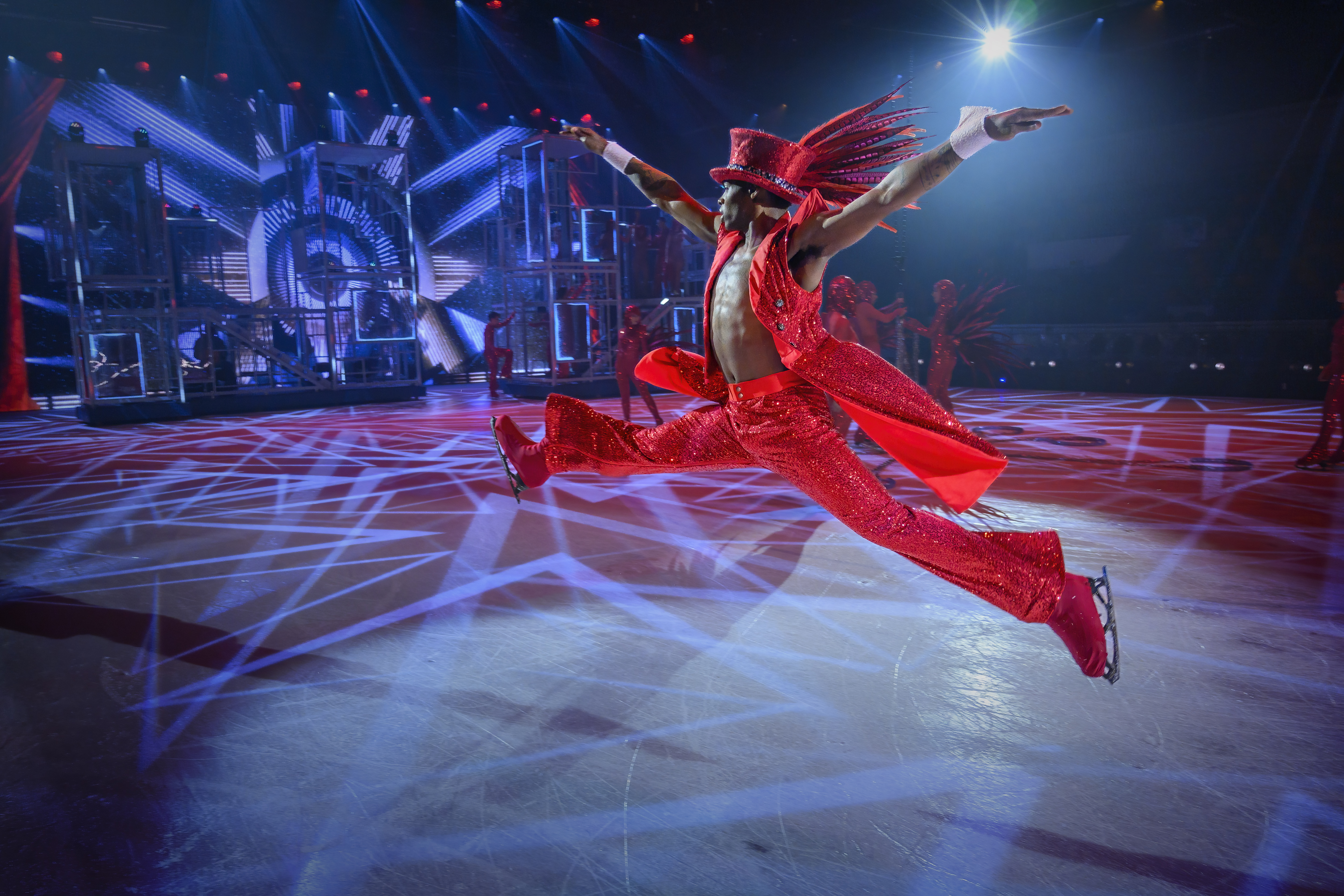 Ein Eiskunstläufer in einem roten, glitzernden Anzug macht einen Sprung mit weit gespreizten Beinen.  