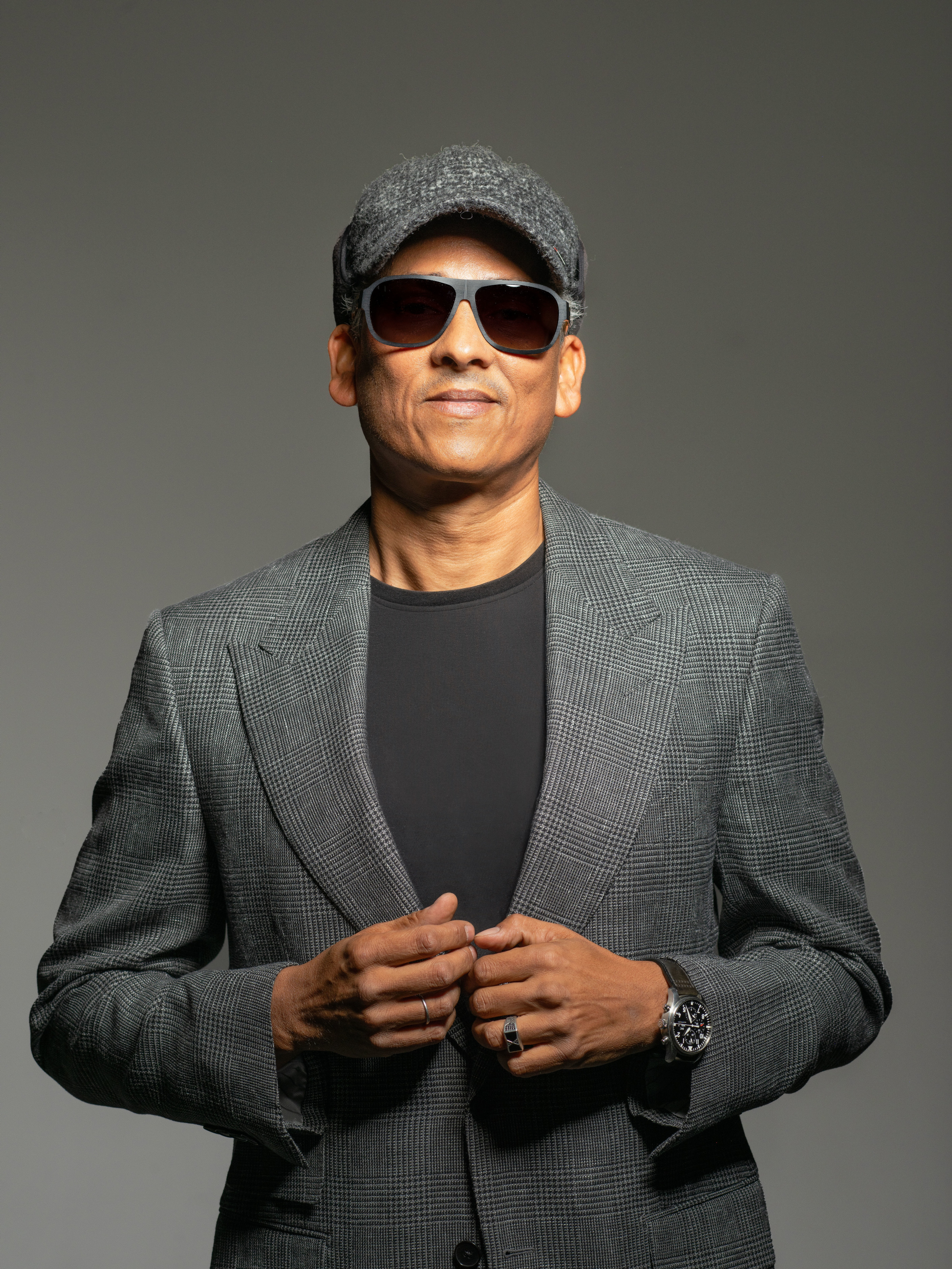 Pressebild des Sängers in Grau- und Brauntönen. Der Sänger trägt eine graue Kappe, eine Sonnenbrille und ein graues Jacket. 